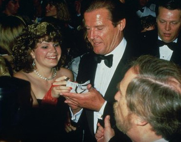Roger Moore signs autographs for the waiting fans as EON Productions’ Director of Marketing Jerry Juroe tries to move him on.